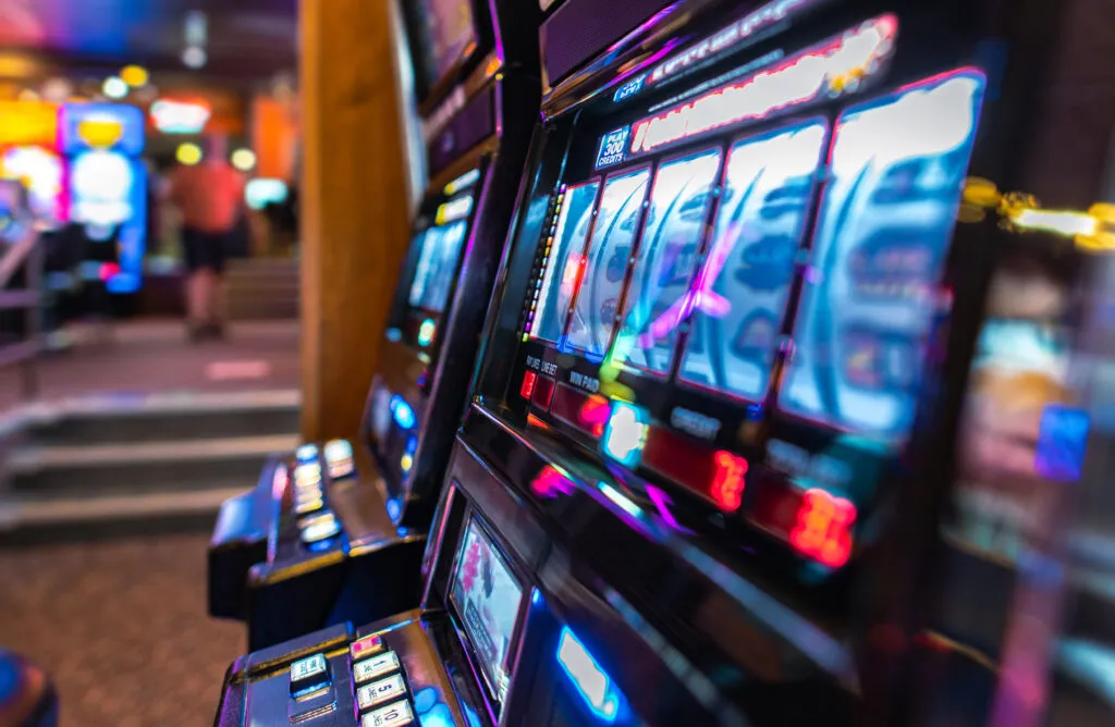 A row of brightly lit slot machines on a casino floor, shot at an angle, with a figure walking away out of focus in the background
